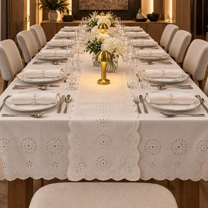 May include: A long rectangular dining table set for a formal meal, featuring a white tablecloth and matching table runner with intricate eyelet embroidery along the scalloped edges. The table is arranged with white plates, napkins, and cutlery.