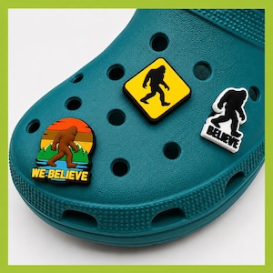 May include: A teal foam clog shoe featuring three Bigfoot-themed charms. One charm is a yellow diamond-shaped sign with a black Bigfoot silhouette, another is a white cutout with a black Bigfoot and the text BELIEVE, and one is a sunset scene.