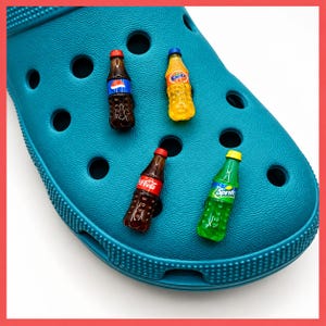 Könnte beinhalten: Ein türkisfarbener Schaumstoff-Clog mit vier Miniatur-Getränkeflaschen-Anhängern, die in die Belüftungslöcher gesteckt sind. Die Anhänger zeigen eine dunkelbraune Pepsi-Flasche, eine orangefarbene Fanta-Flasche, eine Coca-Cola-Flasche und eine grüne Sprite-Flasche.