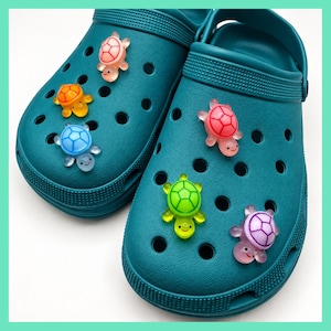 May include: A pair of teal clogs decorated with six small, translucent turtle-shaped charms. The charms are coloured in shades of pink, orange, blue, green, and purple, each featuring a simple, smiling face and a detailed shell pattern on the back.