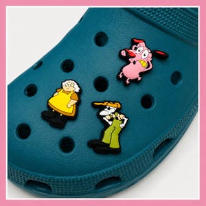 May include: A teal clog shoe featuring three cartoon character charms attached to the ventilation holes. The charms depict a pink dog with wide eyes, an elderly person in a yellow dress, and a thin character wearing green overalls and a white shirt.