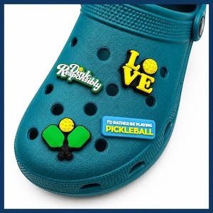 May include: A teal clog shoe featuring four pickleball-themed charms. The charms include text that reads "Dink Responsibly", "LOVE" with a yellow ball, a pair of green pickleball paddles with a yellow ball, and a blue sign saying "I'D RATHER BE PLAYING PICKLEBALL".