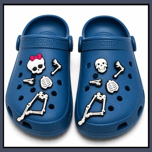 May include: A pair of navy blue clogs featuring white skeleton-themed charms. The left clog displays a skull with a pink bow, while the right features a plain skull. Both shoes include matching white bone charms for the arms, ribs, and legs.