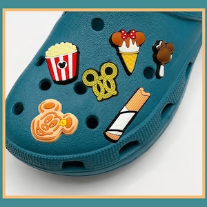 May include: A teal clog shoe decorated with six food-themed charms including a red and white popcorn bucket, a pretzel, a Mickey Mouse-shaped ice cream cone, a chocolate-covered treat, a Mickey-shaped waffle, and a churro-style snack.