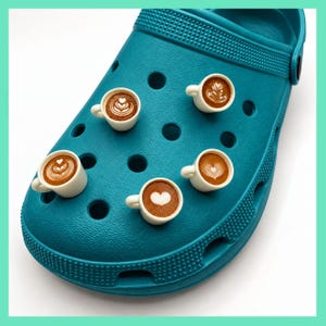 May include: A teal clog shoe featuring five miniature white coffee cup charms attached to the upper surface. Each small cup contains brown liquid with distinct white latte art designs, including heart shapes and intricate rosetta patterns.