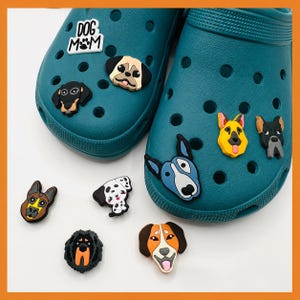 May include: A pair of teal clogs decorated with various rubber dog-themed charms, including a white sign with the text DOG MOM, a pug, a dachshund, a bull terrier, a German shepherd, and a schnauzer, with additional loose dog charms nearby.