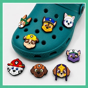 May include: A teal clog shoe featuring several colorful Paw Patrol character charms attached to the ventilation holes. Additional character charms are arranged below the shoe, depicting various dog faces with hats and gear in vibrant colors.