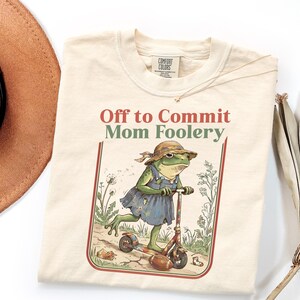 Peut inclure: Un t-shirt couleur crème avec une illustration représentant une grenouille portant un chapeau de soleil et une robe bleue, sur une trottinette. Le texte indique Off to Commit Mom Foolery. Le motif inclut des pissenlits, un ballon et une chaussette.
