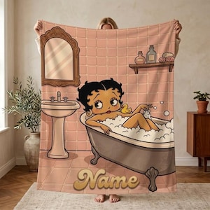 May include: A rectangular fleece blanket featuring a cartoon illustration of a character with black hair in a bathtub filled with bubbles. The scene includes a pedestal sink, a wall mirror, a shelf with bottles, and the word Name in gold script.
