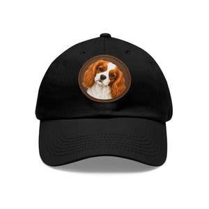 Op de afbeelding: Een zwarte baseballpet met een ronde patch op de voorkant. De patch toont een gedetailleerde illustratie van een Cavalier King Charles-spaniël met lang, golvend kastanjebruin en wit haar, een zwarte neus en grote bruine ogen, met een bruine rand.