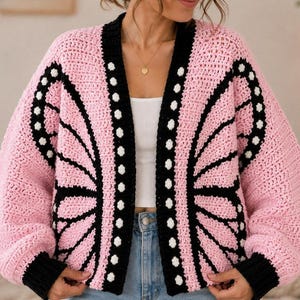 May include: A crochet cardigan featuring a pink base with black and white butterfly wing patterns. The garment has long sleeves with black ribbed cuffs, a black front border with white circular accents, and an open front design.