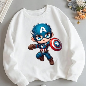 Puede incluir: Una sudadera blanca de manga larga con un gr&aacute;fico central de un personaje de dibujos animados de Capit&aacute;n Am&eacute;rica. El personaje lleva una m&aacute;scara azul con una A blanca, un traje azul y rojo con una estrella blanca y sostiene un escudo circular.