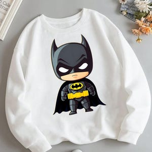 Puede incluir: Una sudadera blanca de manga larga con un estampado gr&aacute;fico de un personaje de Batman al estilo chibi. El personaje viste un traje negro, una capa negra y un cintur&oacute;n utilitario amarillo, con el ic&oacute;nico s&iacute;mbolo de murci&eacute;lago amarillo y negro en el pecho.