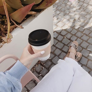 Coffee cup with a sticker held by a hand. Italian vegetable-tanned leather coffee cozy for ultimate comfort. Personalized and adjustable to fit all cup sizes