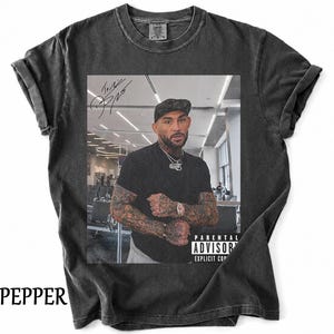 May include: A charcoal grey short-sleeve t-shirt featuring a graphic print of a tattooed individual wearing a black cap and t-shirt. The print includes a signature in the top left and a black and white Parental Advisory Explicit Content label.