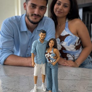 Op de afbeelding: Een op maat gemaakt beeldje van een koppel dat samen op een ronde witte voet staat. Het mannelijke figuur draagt een lichtblauw overhemd, witte korte broek en witte sneakers. Het vrouwelijke figuur draagt een blauw-wit patroon haltertop en een spijkerbroek.