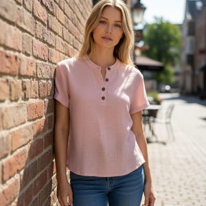 May include: A light pink short-sleeved shirt with a mandarin collar and a partial button-down front featuring four dark brown buttons. The shirt has a textured fabric finish and is paired with blue denim jeans, creating a casual look.