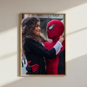 Op de afbeelding: Een ingelijste fotoprint met een persoon met lang golvend bruin haar in een zwart jasje en rode handschoenen, die een figuur in een rood en zwart Spider-Man-pak met witte ooglenzen omhelst. De afbeelding zit in een lichthouten lijst.