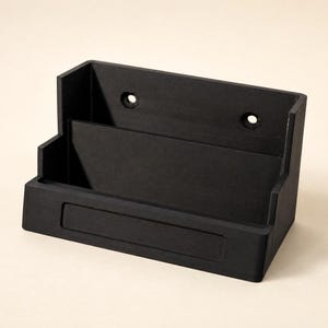 May include: A matte black wall-mounted organizer featuring a tiered design with two distinct compartments for storage. The back panel includes two circular holes for mounting, and the front face displays a rectangular recessed area.