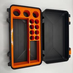 Puede incluir: Un estuche de almacenamiento rectangular negro con la tapa abierta, que presenta un inserto interior de color naranja brillante. El inserto contiene tres ranuras circulares en la parte superior, ocho ranuras circulares pequeñas y dos compartimentos.