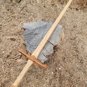 May include: A handcrafted wooden sword featuring a long, light-colored blade with a pointed tip. The crossguard and pommel are constructed from a darker, contrasting wood with visible grain patterns, attached to a smooth, cylindrical handle.