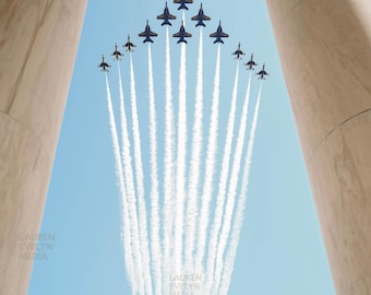 May include: A formation of eleven dark blue fighter jets flying in a V-shape against a clear light blue sky, leaving long white vapor trails behind them, framed by two large, smooth, light-colored stone columns on the left and right sides.