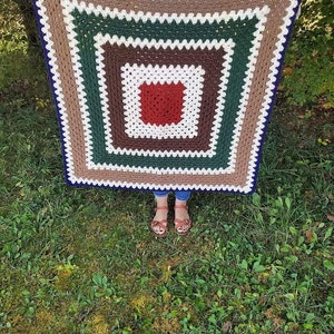 May include: A crocheted square blanket with a multi-colored border and a red center square. The blanket is being held by a person in a grassy area.