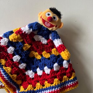 May include: A handmade crochet security blanket featuring a plush character head. The blanket is a patchwork of red, white, blue, and yellow yarn. The character has a yellow face, black hair, and a red nose and mouth.