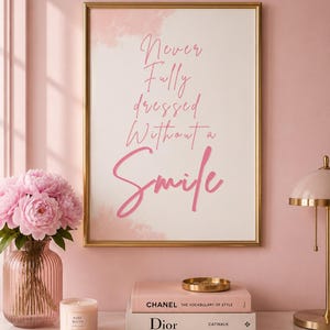 Puede incluir: Una lámina decorativa rectangular en un marco dorado con el texto en rosa Never Fully dressed Without a Smile sobre un fondo blanco con suaves toques de acuarela rosa. La lámina se muestra en una habitación con paredes y decoración rosa.