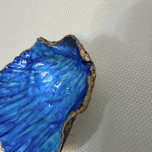 Könnte beinhalten: Eine Muschelschale mit einem leuchtend blauen Inneren, das strukturierte Pinselstriche und einen goldfarbenen Rand aufweist. Die Muschel hat eine unregelmäßige, organische Form mit einer glänzenden Oberfläche, die blaue und cyanfarbene Töne betont.
