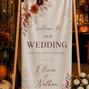 May include: A vertical fabric wedding sign on a wooden easel featuring a beige linen texture, decorated with watercolor floral arrangements in burgundy, rust, and cream. The text reads: welcome to OUR WEDDING WE'RE SO GLAD YOU'RE HERE Olivia & Nathan OCTOBER 20, 2026.