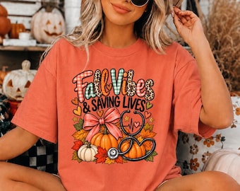 May include: A coral-colored t-shirt featuring a graphic print with the text Fall Vibes & SAVING LIVES. The design includes pumpkins, a pink bow, a stethoscope, and autumn leaves in shades of orange, yellow, and green with plaid patterns.