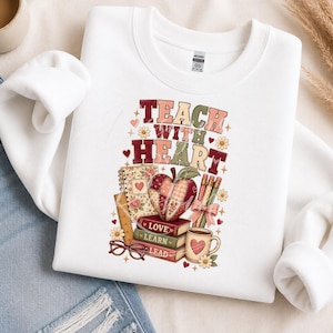 Pode incluir: Uma camisola branca de gola redonda com uma impressão gráfica com o texto "TEACH WITH HEART" numa fonte multicolorida. O design inclui uma maçã com padrão, uma pilha de três livros com as etiquetas "LOVE", "LEARN", "LEAD", lápis e flores.