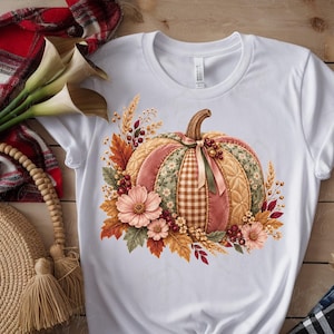 May include: A white short-sleeved t-shirt featuring a central graphic of a pumpkin with alternating panels of plaid, floral, and quilted patterns in muted autumn tones, accented with pink flowers, wheat stalks, and small red berries.