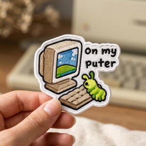 May include: An embroidered patch featuring a beige vintage computer with a blue sky and green hill screen display, a brown keyboard, and a small green caterpillar. The text "on my puter" is embroidered in black next to the computer.