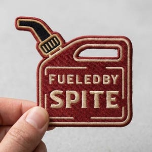 May include: A red embroidered patch shaped like a fuel can with a black nozzle. The patch features the text FUELED BY SPITE in beige capital letters, enclosed within a beige rectangular border. The design is stitched with a dark red border.