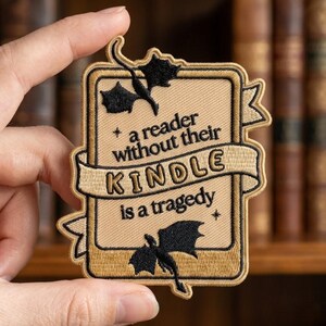 May include: A rectangular embroidered patch with a tan background and dark brown border. It features two black dragon silhouettes and the text: a reader without their KINDLE is a tragedy. The design includes a central banner across the middle.