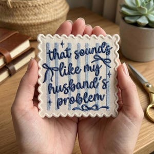 May include: A square fabric patch with scalloped edges featuring a light blue and white vertical striped pattern. The text "that sounds like my husband's problem" is embroidered in dark blue cursive script, accented by small embroidered stars and bows.