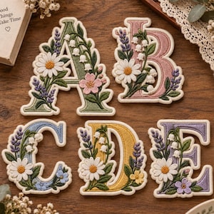 May include: Five embroidered fabric letters A, B, C, D, and E, each featuring a unique pastel color and detailed floral motifs including white daisies, lavender sprigs, and lily of the valley, with a cream-colored border around each letter.