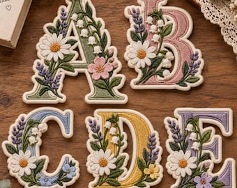 May include: Five embroidered fabric letters A, B, C, D, and E, each featuring a unique pastel color and detailed floral motifs including white daisies, lavender sprigs, and lily of the valley, with a cream-colored border around each letter.