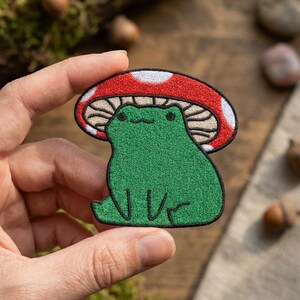 May include: An embroidered patch featuring a bright green frog sitting under a large red mushroom cap with white spots. The frog has a simple black stitched face with two dots for eyes and a small curved line for a mouth. The entire design is outlined in black thread.