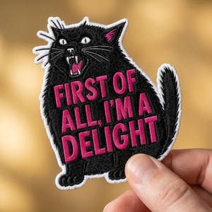 May include: An embroidered patch shaped like a black cat with white whiskers, pink inner ears, and an open mouth showing teeth. The text FIRST OF ALL, I'M A DELIGHT is stitched in bold pink capital letters across the center of the cat's body.