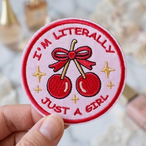 May include: A circular embroidered patch with a light pink background and a red border. It features two red cherries tied with a red bow, surrounded by gold star accents. The text "I'M LITERALLY JUST A GIRL" is embroidered in red around the cherries.
