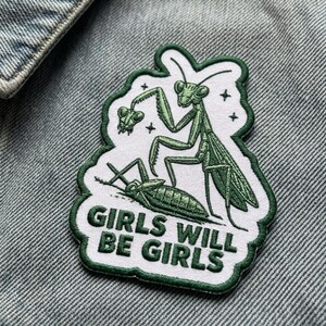May include: An embroidered patch with a white background and a dark green border featuring a green praying mantis holding a smaller insect. The text GIRLS WILL BE GIRLS is embroidered in dark green capital letters at the bottom of the patch.