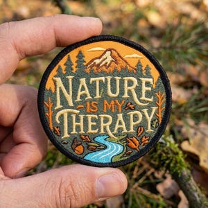May include: A circular embroidered patch featuring a mountain landscape with a blue river, green pine trees, and orange sky. The text "NATURE IS MY THERAPY" is stitched in cream-colored lettering across the center, framed by a black border.