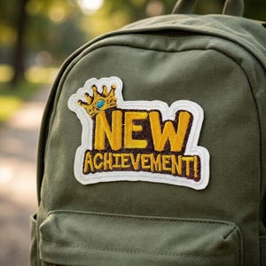May include: An olive green canvas backpack featuring an embroidered patch with a white border. The patch displays a gold crown with a blue gem above the yellow text NEW ACHIEVEMENT! set against a dark brown background on the fabric surface.