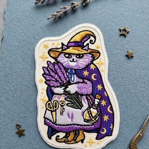 May include: An embroidered patch featuring a purple cat wearing a yellow witch hat and a purple cape with gold stars and moons. The cat holds a bundle of lavender and wears an apron with a pair of scissors, a key, and a small glass bottle.
