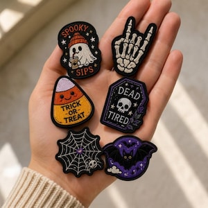 May include: Six embroidered patches featuring Halloween themes: a ghost in a beanie with the text SPOOKY SIPS, a skeleton hand, a smiling candy corn with TRICK-OR-TREAT, a coffin with a skull and DEAD TIRED, a spider web with a spider, and a bat.