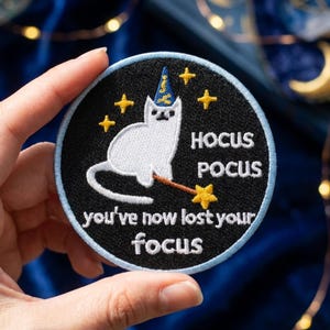 May include: A circular embroidered patch with a black background and a light blue border. It features a white cat wearing a blue wizard hat, holding a wand with a yellow star, surrounded by yellow stars, with the text HOCUS POCUS you've now lost your focus.