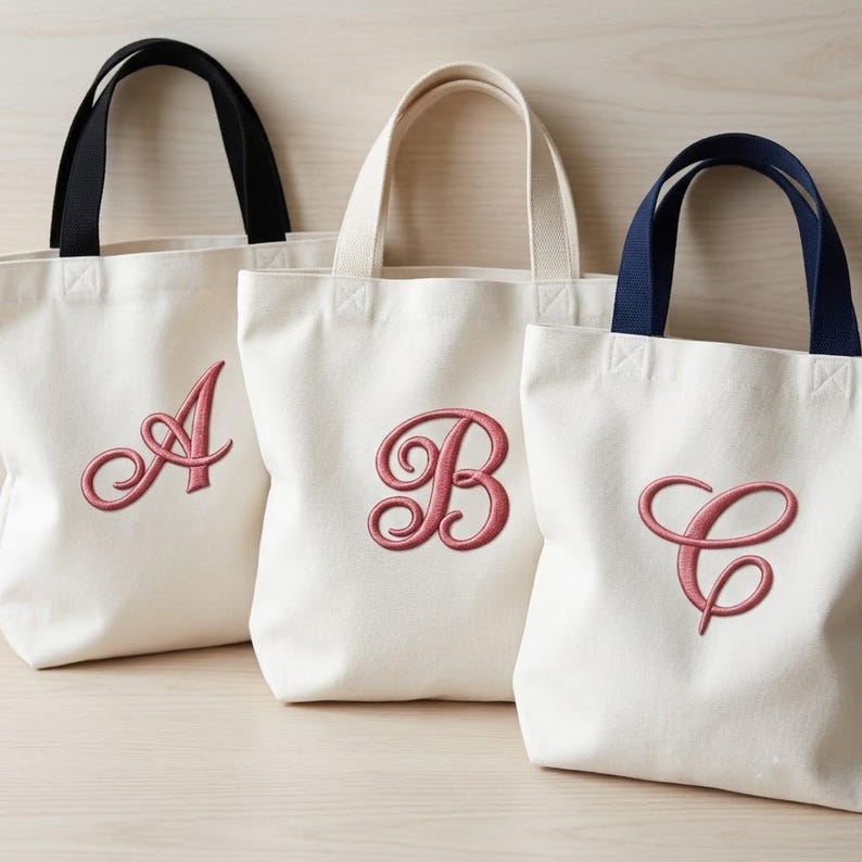 K&ouml;nnte beinhalten: Drei cremefarbene Canvas-Tragetaschen mit jeweils zwei Henkeln. Die Taschen sind mit den gestickten Schreibschriftbuchstaben A, B und C in Altrosa versehen. Die erste Tasche hat schwarze Henkel, die zweite cremefarbene und die dritte dunkelblaue Henkel.